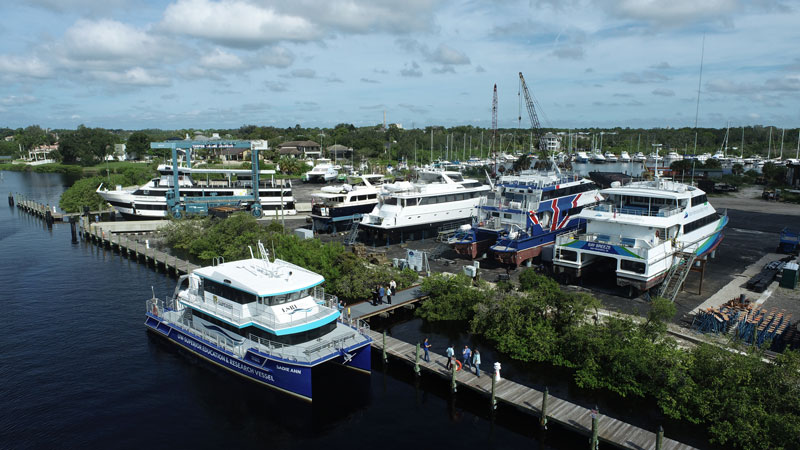 The Sadie Ann research vessel is docked among yachts at the tree-lined marina shore.