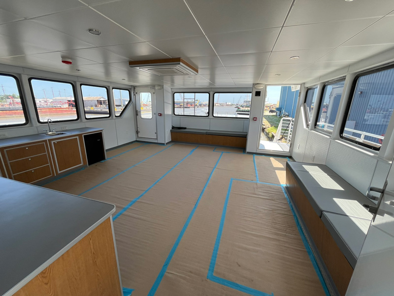 Spacious room with large windows, wood cabinets, and counters. The floor is covered with protective cardboard taped down. The area appears to be an unfurnished interior of a boat or ferry, with the dockyard visible about the windows outside.