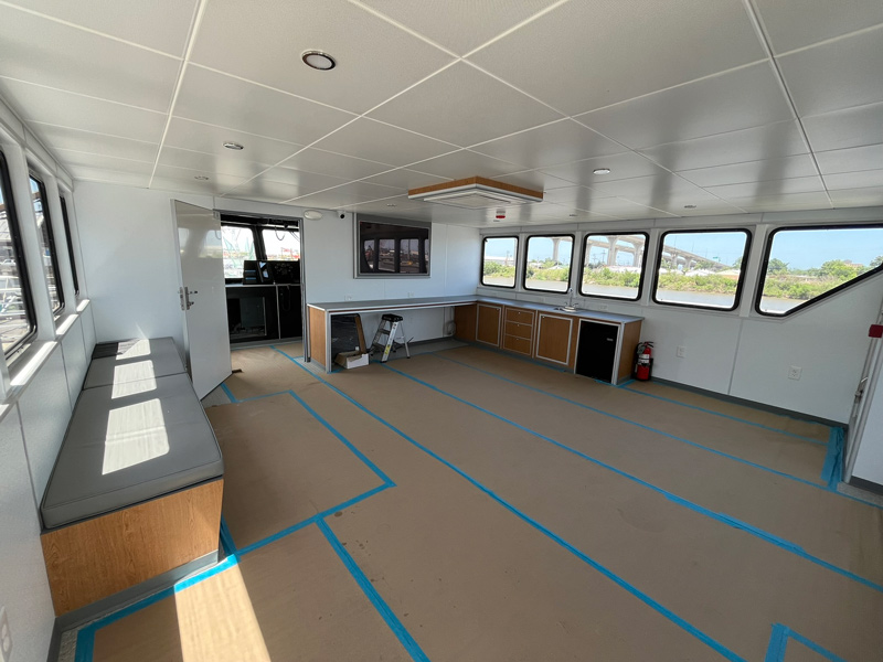 Spacious, modern room with large windows, a built-in bench along one wall, and blue tape marking areas on the floor about the space. Sunlight streams in, highlighting a counter, cabinets, and a fire extinguisher in the corner.