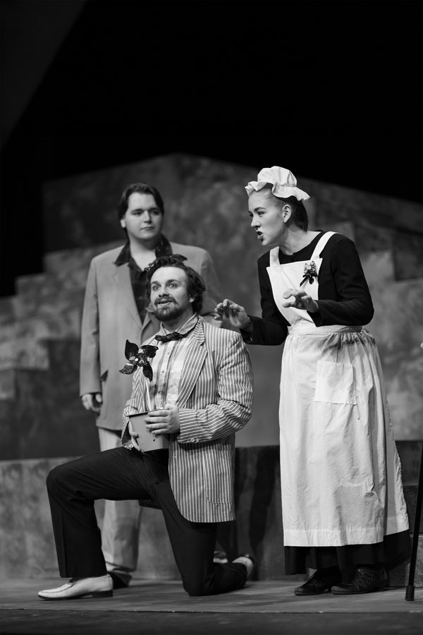 Three actors on stage in a theatre; one kneels with a flowerpot, another stands, one’s a maid.