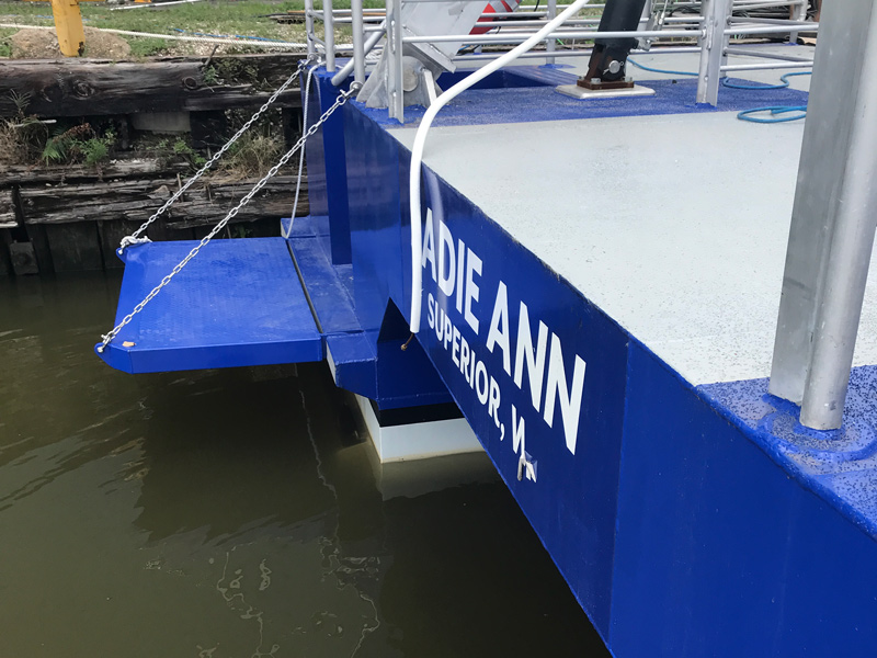A blue boat named “Sadie Ann” with “Superior, WI” written below on its side is docked about a small blue boarding platform with a metal chain that hangs over the water nearby.