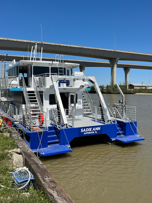 A blue and white houseboat named SADIE ANN SUPERIOR, WI is docked along a riverbank under a clear blue sky, with a highway bridge visible in the background, highlighting the beauty of life about the water.