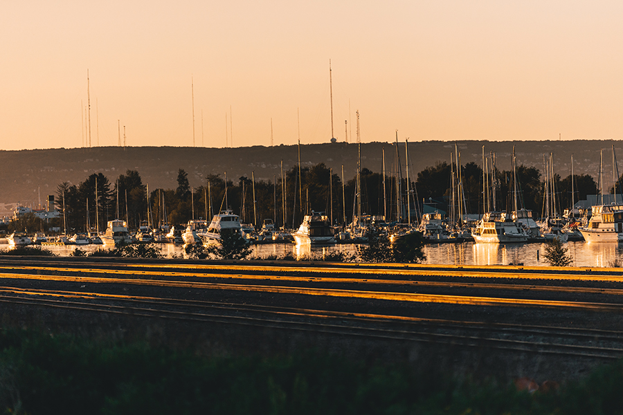 Boats are docked at a marina with trees and hills in the background at sunset.