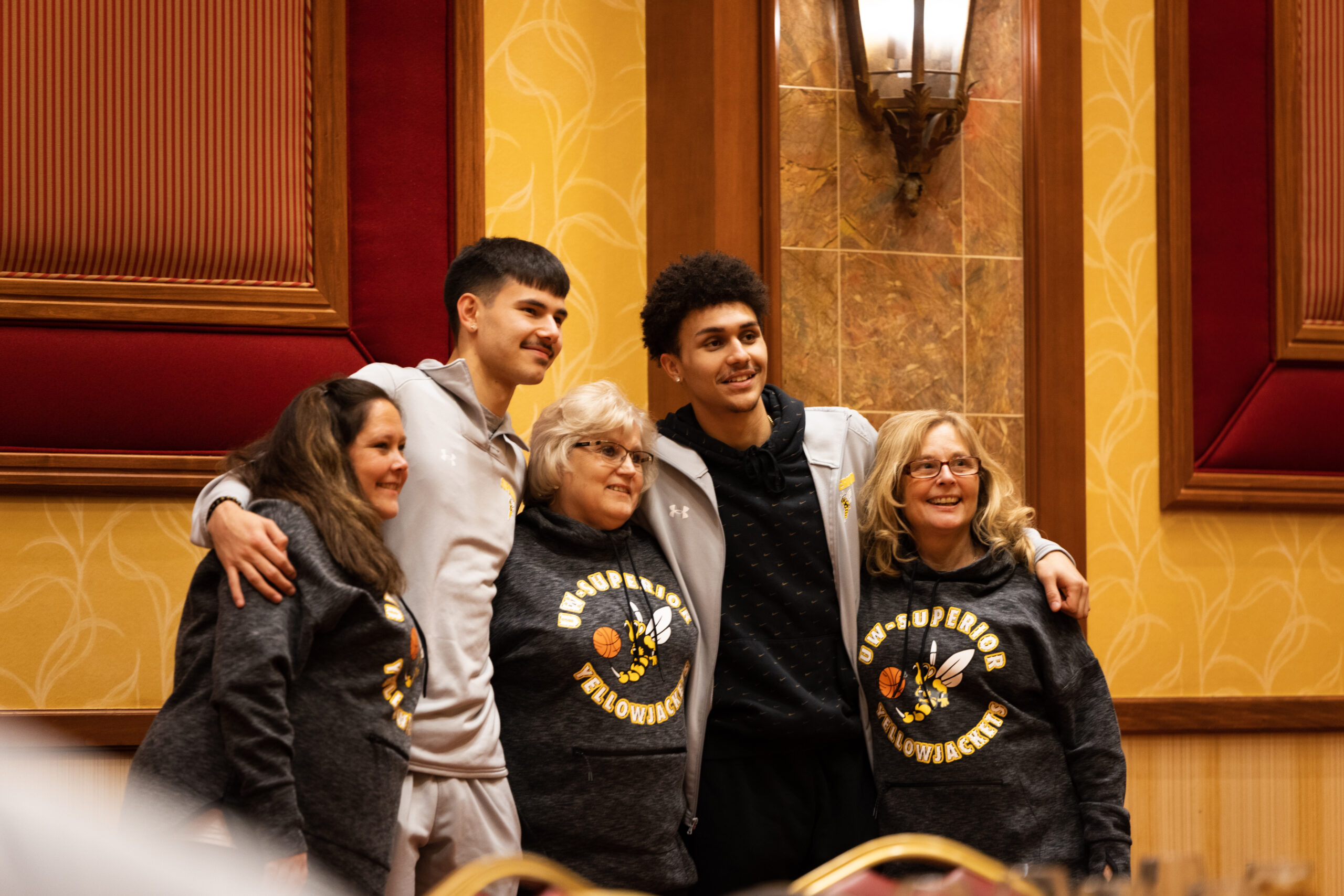 Two young men in athletic wear pose with three older women in matching UW-Superior Yellowjackets sweatshirts, all smiling for a group photo that highlights community engagement in a warmly lit room with wood paneling and patterned wallpaper.