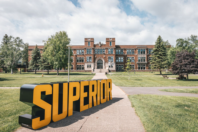Large SUPERIOR sign stands before a brick academic building, symbolizing graduation success.