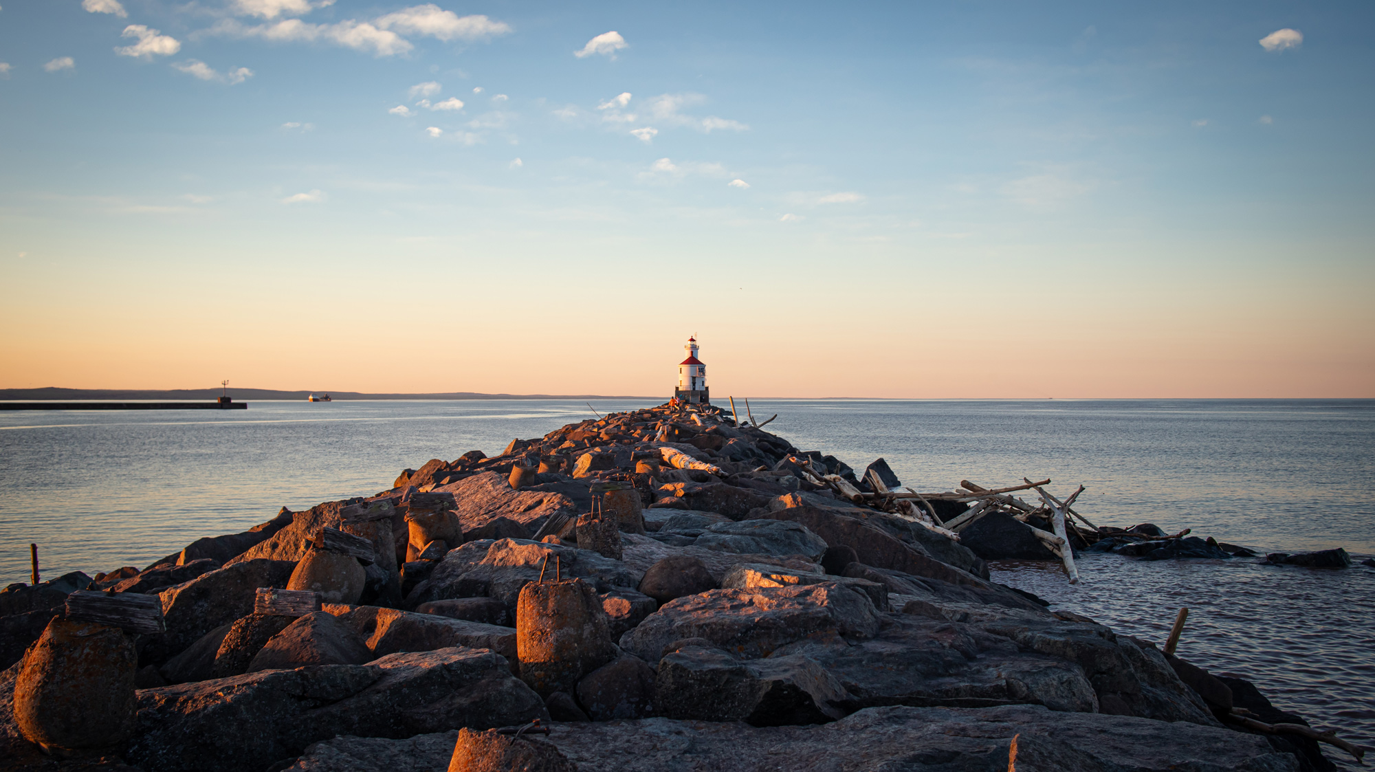 A small lighthouse sits at the end of a rocky pier, stretching into calm water under a clear sky at sunset. Driftwood is scattered about the rocks, and distant land is visible on the horizon.
