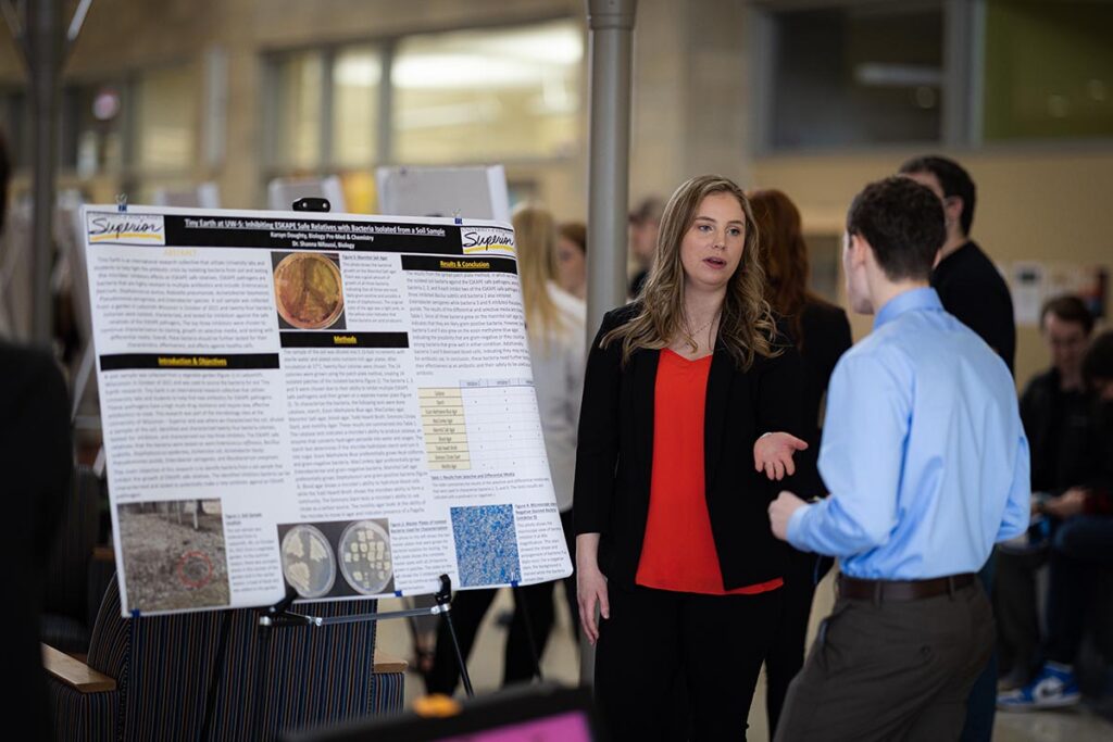 A woman and a man stand in front of a Natural Sciences poster at a conference or academic event, engaged in conversation. Other people are visible in the blurred background.