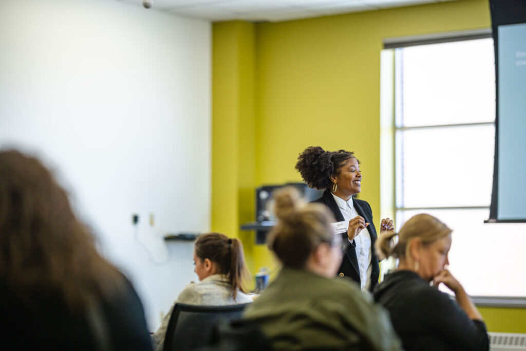 A woman stands and smiles while presenting to a group of seated people in a bright, modern classroom with a yellow wall and large window, leading a Childcare Center Administration workshop.