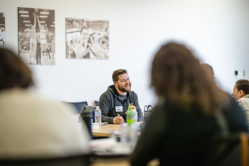 A man sits at a table, smiling and talking with others during a meeting on Learning Strategies for Educators. Several people are seated around him, while black-and-white sports photos hang on the white wall in the background.