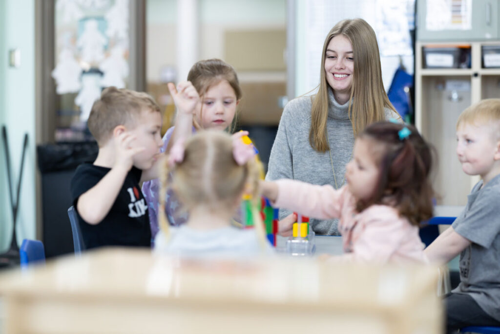 A teacher sits at a table with five young children playing with colorful building blocks in a classroom, all appearing engaged and happy—showcasing an inviting environment supported by effective Childcare Center Administration.