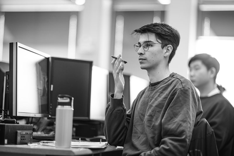 Person sits at a computer desk holding a pen, focusing on cybersecurity, another in the background.