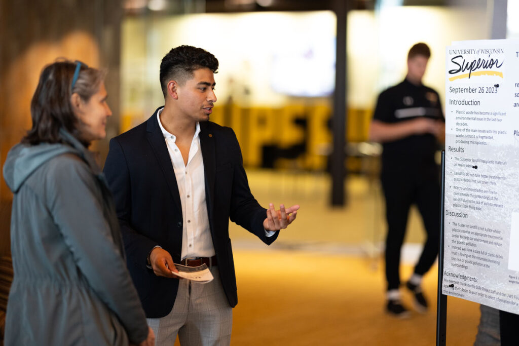 A man in a suit explains information about a poster to a woman at an indoor event; another person is blurred in the background. The poster displays text and the University of Wisconsin-Superior logo.