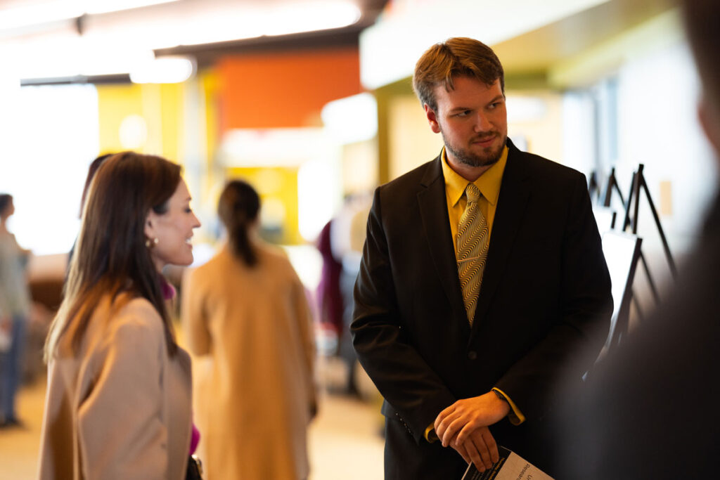 A man in a suit and yellow shirt stands holding papers, talking to a woman with brown hair and a light coat about something. Other people and blurred colorful backgrounds are visible indoors.