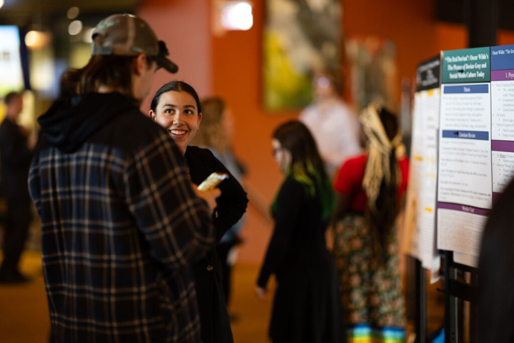 Two people stand and talk about a presentation board at an indoor event, while others in the background look at posters. The setting is casual and brightly lit.