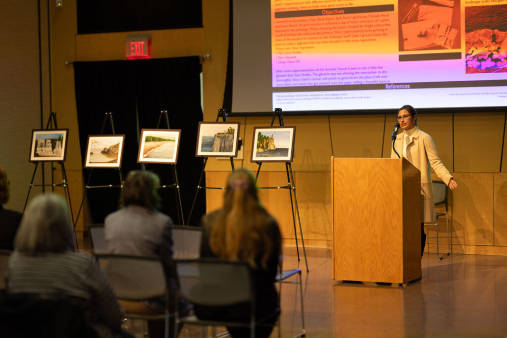 A woman stands at a podium giving a presentation to a small seated audience about her topic. Behind her are large display boards with images on easels and a projection screen with text and photos in an indoor event or lecture setting.