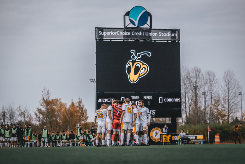 Men's Soccer in a huddle below the big screen displaying yellowjacket mascot Buzz.