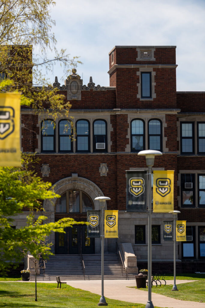 Old Main building with arched entrance showing yellow UWS banners on campus green lawn.
