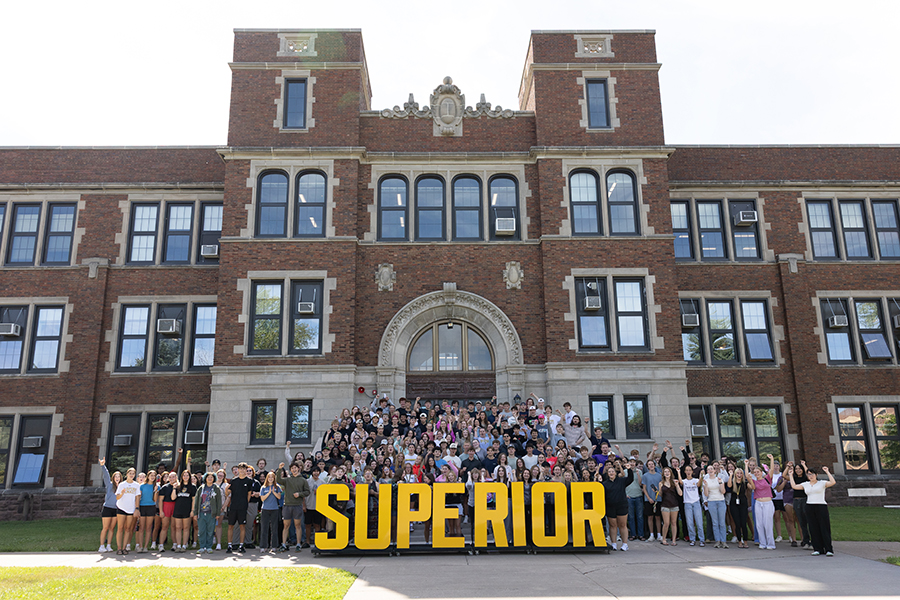A large group stands in front of a brick building holding large yellow letters spelling SUPERIOR..