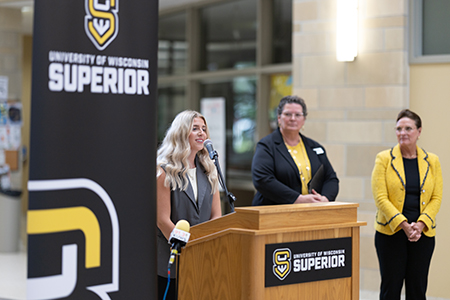 A woman speaks at a podium with two women standing behind her at University of Wisconsin-Superior.