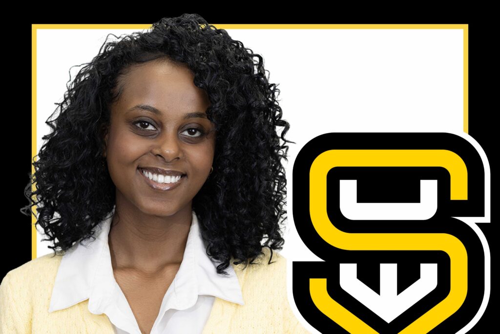 Woman with curly hair smiling, wearing a white shirt and yellow sweater, with bold logo.