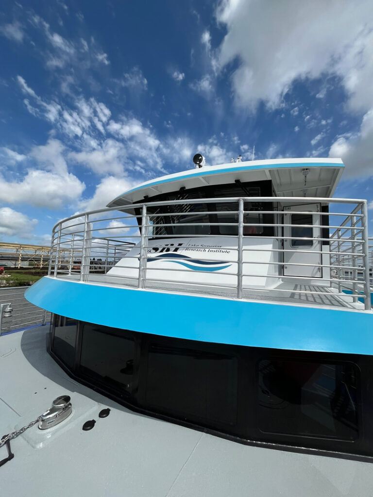 A close-up view of a boat’s upper deck and cabin with blue and white colors, metal railings, and partly cloudy skies above tells about the Louisiana Universities Marine Consortium, as seen on the vessel’s side.