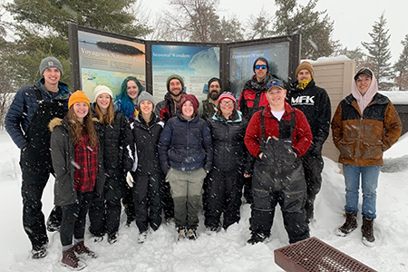 A group of people in winter clothing poses for a photo outside in the snow by informational signs.