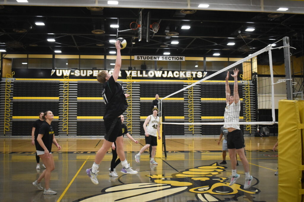 A group of people play indoor volleyball; one player in black jumps to spike the ball over the net while an opponent in white tries to block. The gym has UW-Superior Yellowjackets signs and a large yellow jacket logo on the floor.