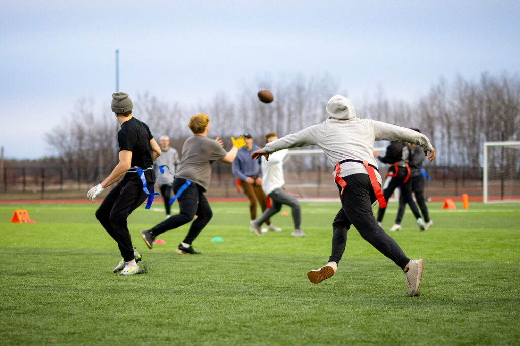 A group of people playing flag football outdoors on a grassy field. One person in a gray hoodie throws a football while others run and reach for the ball, with trees and goalposts visible in the background.