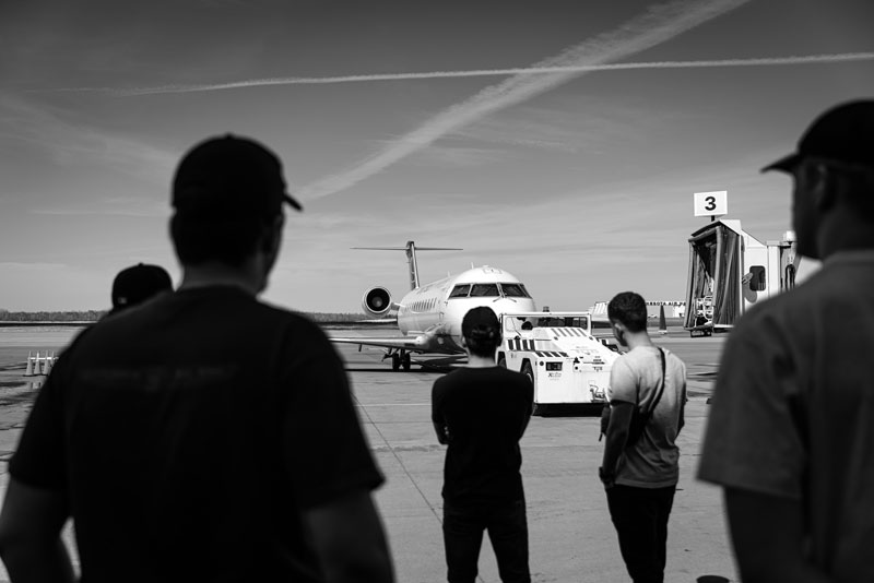 Four people watch a jet being towed, discussing airport economics near the boarding gate.