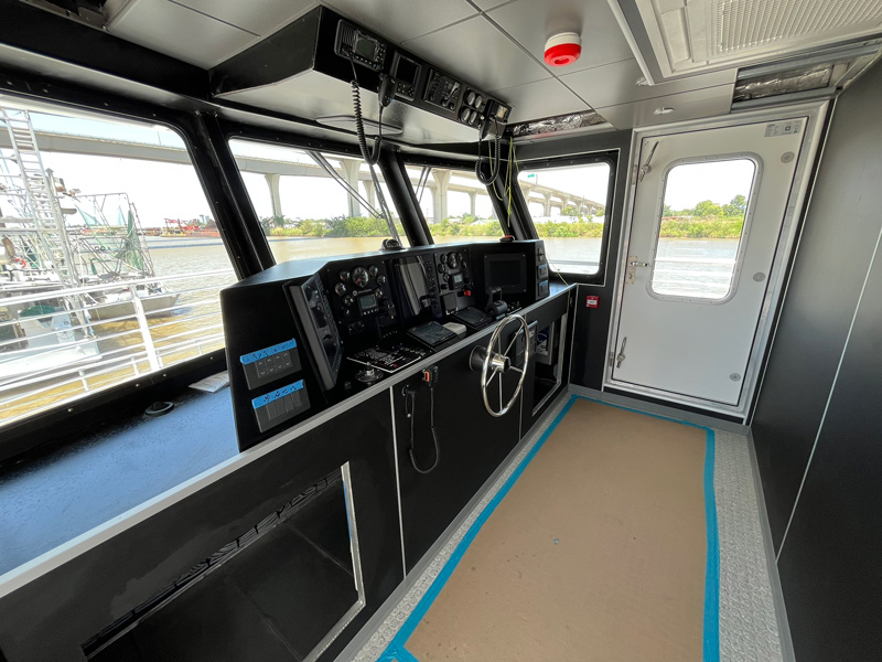 Interior of a modern boat’s control room with steering wheel, control panels, and electronic displays. Large windows offer views of the river, bridge, and shoreline. A tan mat with blue tape covers the floor, adding detail to this space all about navigation.