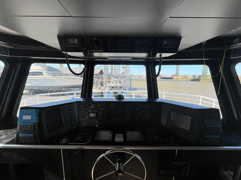 View from inside a boats cockpit, showing navigation controls, instruments, and steering wheel, with large windows overlooking a marina—about as close as you can get to being at the helm, surrounded by boats and water outside.
