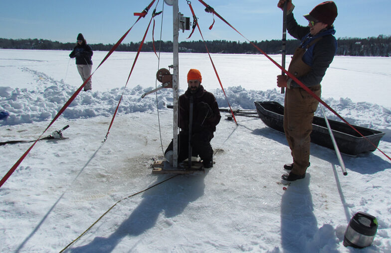 Three people work with equipment on a frozen lake, drilling through the ice.