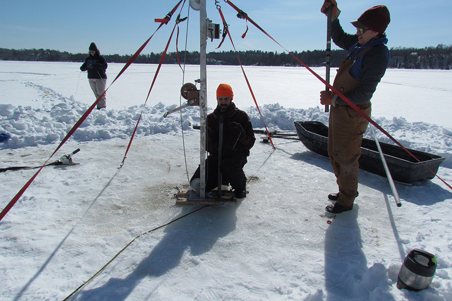 Three people work with equipment on a frozen lake, drilling through the ice.