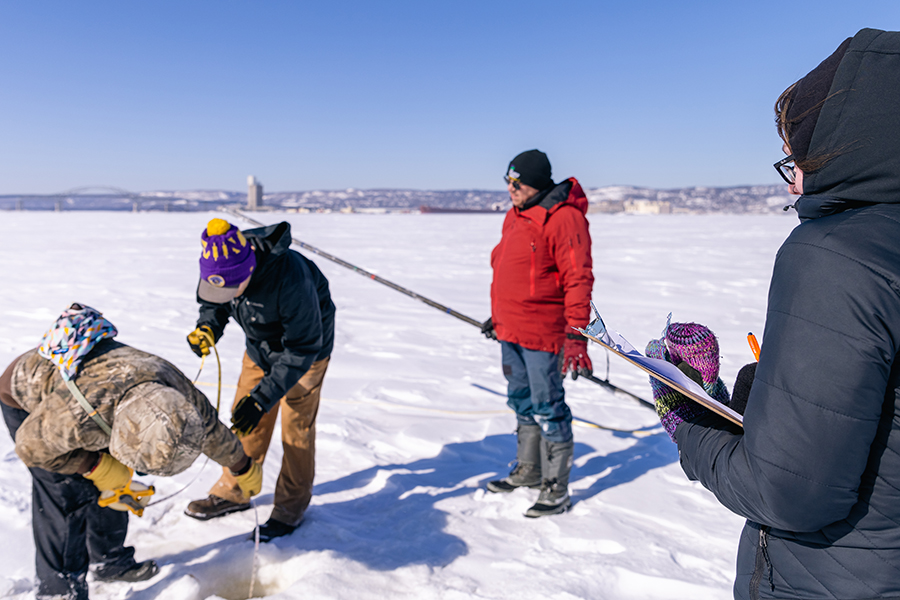 Four people gather on a snowy field, drilling into ice and taking notes on a clipboard.