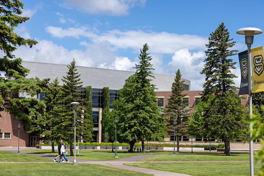 A campus building with large windows, trees, a lawn, and two people walking on a path.