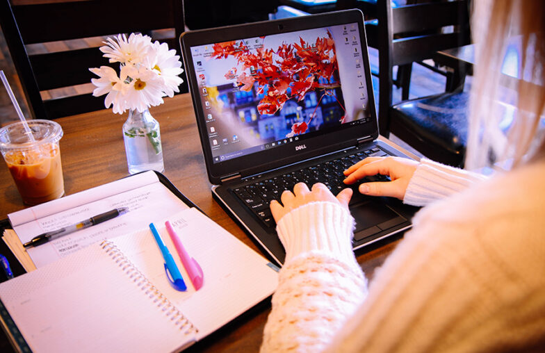 Person typing on a laptop at a table with notebooks, pens, iced coffee, and a vase of flowers.