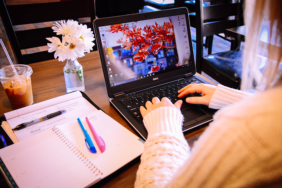 Person typing on a laptop at a table with notebooks, pens, iced coffee, and a vase of flowers.