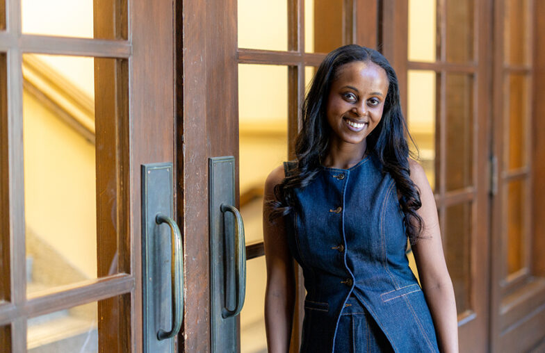 Woman in a sleeveless denim dress standing and smiling by wooden glass doors indoors.