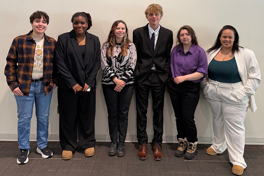 Six people stand in a row against a plain wall, dressed in a mix of casual and formal clothing.