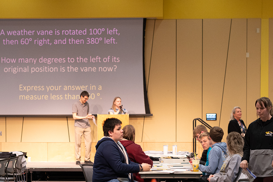 A group of people sit at tables in a large room, while two people stand at a podium in front of a projected math problem about a weather vane. Several others stand nearby, and the room has yellow walls.