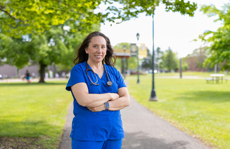 A person in blue scrubs stands on a path in a park, arms crossed, with a stethoscope.
