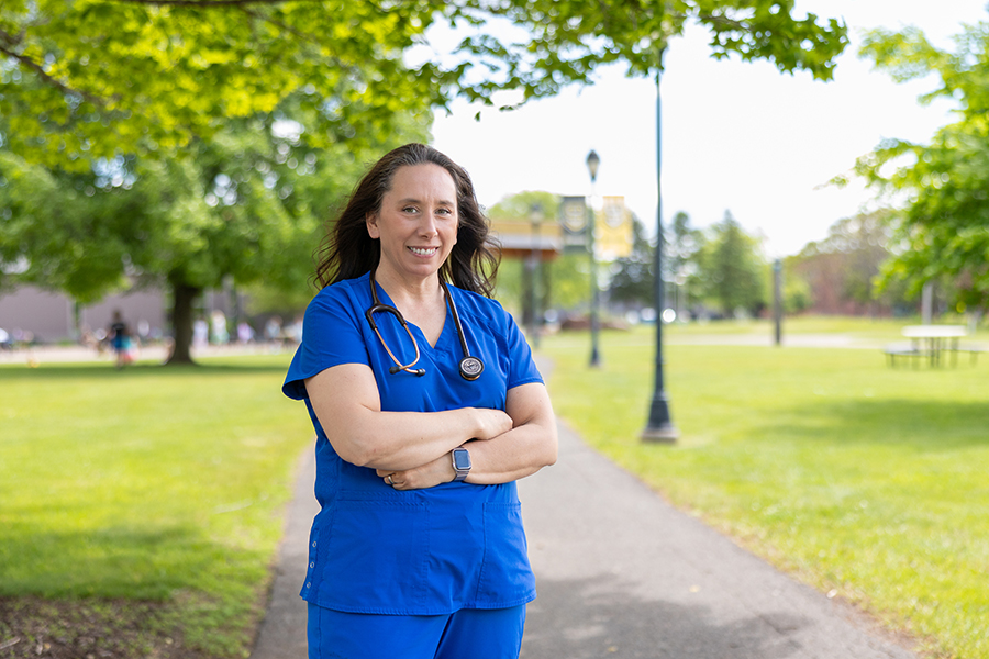 A person in blue scrubs stands on a path in a park, arms crossed, with a stethoscope.