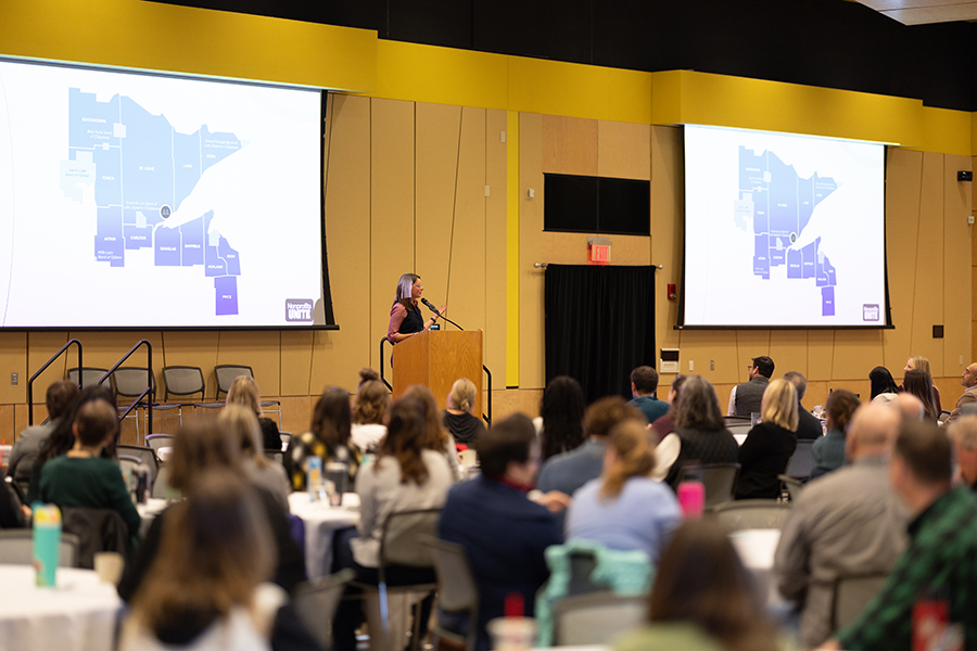 A speaker presents a map slideshow to an audience in a large conference room.