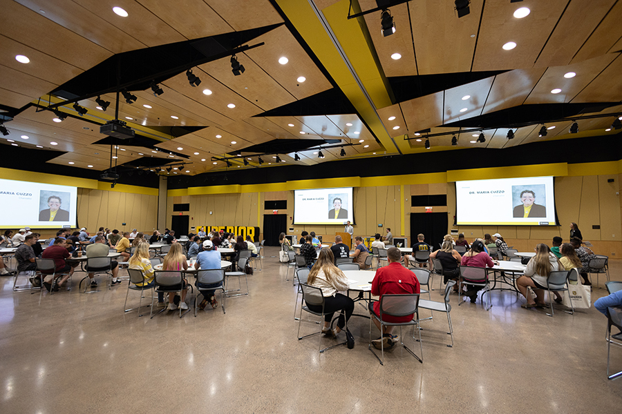 People sit at tables in a large hall, watching a presentation displayed on multiple screens.