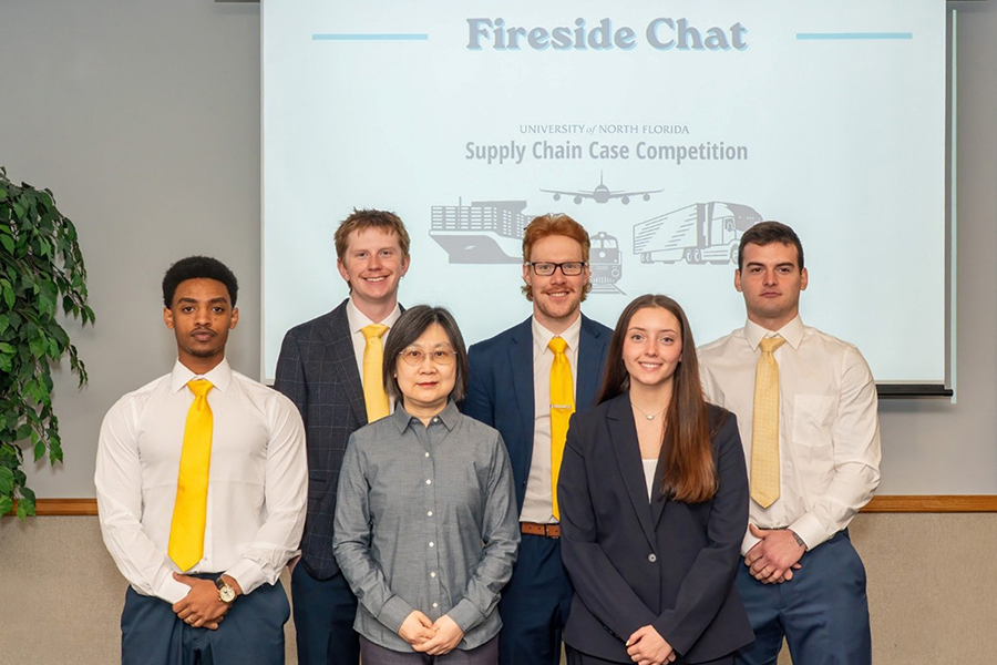 Six people stand in front of a screen displaying “Fireside Chat” and “Supply Chain Case Competition.” Five wear business attire with yellow ties, one wears a gray shirt. They pose and smile for the photo.