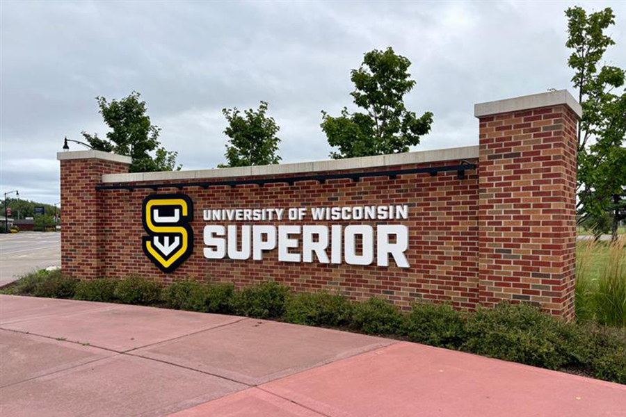 A brick sign with the University of Wisconsin–Superior logo and large white text reading UNIVERSITY OF WISCONSIN SUPERIOR stands next to a sidewalk, with trees and a cloudy sky in the background.