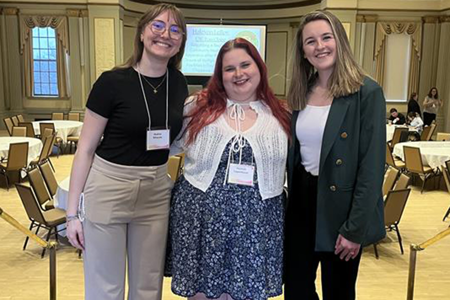 Three people stand in a banquet hall, smiling at the camera, with tables and chairs behind them.
