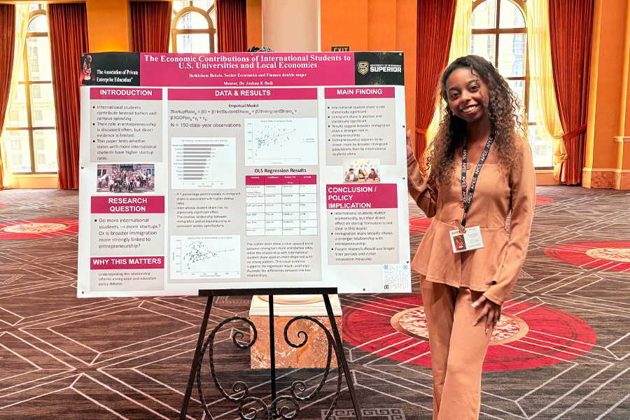 A woman in a tan outfit stands smiling next to a research poster titled The Economic Contributions of International Students to U.S. Universities and Local Economies in a conference room with large windows and patterned carpet.