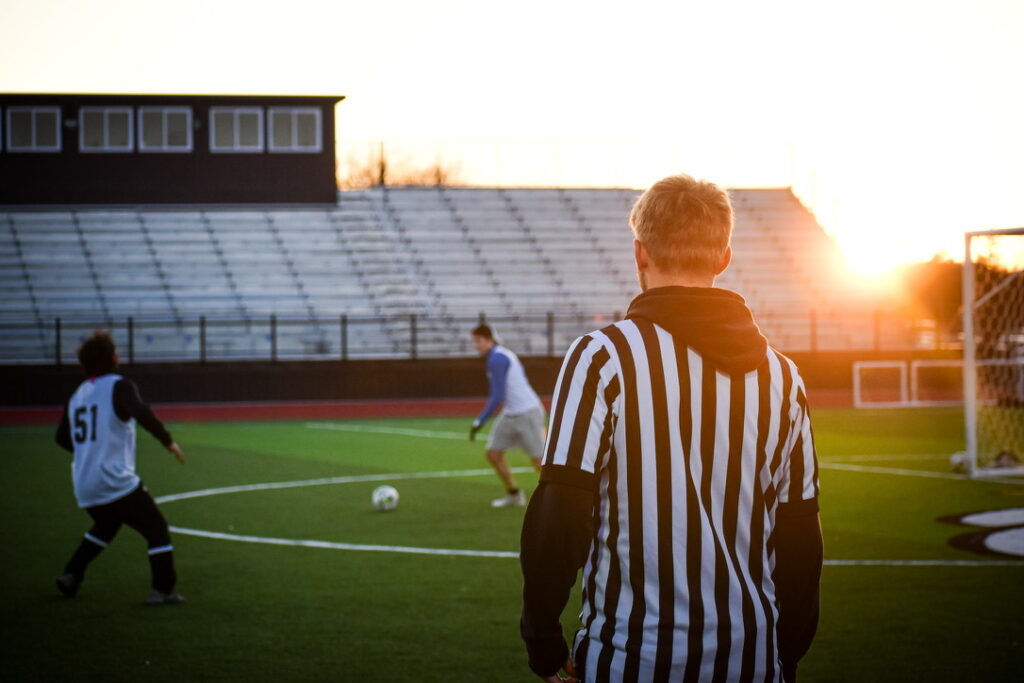 A referee watches two soccer players on a field during sunset, with empty bleachers in the background and the sun low in the sky.