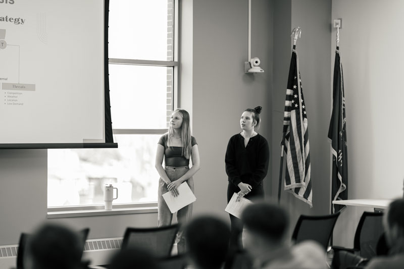 Two people discuss marketing by a projector screen during a presentation in a room with flags.